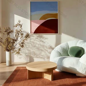 Puede incluir: Un salón moderno con una mesa de centro redonda de madera, una alfombra de color óxido y un sofá blanco curvado con un cojín verde. Una obra de arte abstracta enmarcada cuelga de la pared, con colores azul, rosa y marrón.