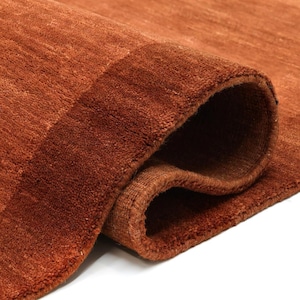 May include: Close-up of a rolled-up, rust-colored rug. The rug has a textured surface with a darker border. The fibers are visible, creating a soft, inviting appearance. The rug's warm tones suggest a cozy and comfortable home decor item.