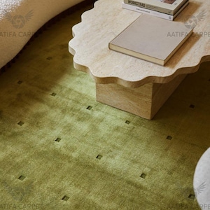 May include: A beige travertine coffee table with a scalloped edge and a rectangular base, topped with two books. One book is titled WORKSTEAD. The table sits on a textured olive green rug featuring a grid of small, recessed square patterns.