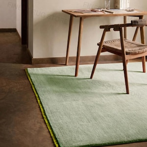 May include: A pale green rug with a vibrant yellow border sits on a brown floor. A wooden desk and chair are in the background. The desk has books and stationery on top. The chair has a woven seat.