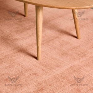 May include: A light brown wooden coffee table with three tapered legs stands on a textured, solid dusty pink carpet. The table features a smooth, oval-shaped top surface and a light wood grain finish, providing a clear view of the furniture base.