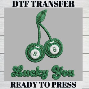 Peut inclure: Un motif de transfert DTF avec deux cerises vertes, chacune portant le numéro 8, et les mots "Lucky You" dans une police verte en forme de corde. Le texte "READY TO PRESS" est en bas, avec "DTF TRANSFER" en haut.