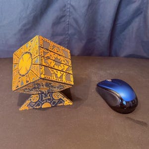 May include: A gold and black puzzle box with intricate geometric designs, resting on a matching stand. A blue computer mouse is next to the puzzle box. The puzzle box is a cube shape.