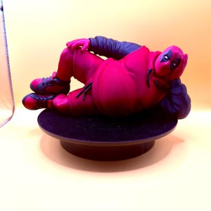 May include: A figurine of a character wearing a red and black hooded sweatshirt and matching shorts, lying in a reclining pose. The character has a red mask with black eye patches, black sleeves, and red and black sneakers, resting on a black circular base.