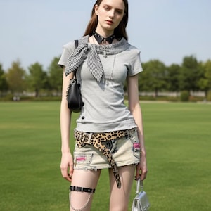 May include: A woman wearing a gray t-shirt, denim mini-skirt, and a leopard print belt. Accessories include a black choker, a black purse, a silver purse, and a black garter with a chain. The outfit is styled with a black and white scarf.