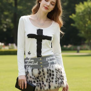 May include: A woman's outfit featuring a white long-sleeve shirt with a large black cross graphic. The outfit includes a gray and leopard print skirt, and a black clutch. The shirt has the text "condus understood like" and "ppines sunge ling" on the skirt.