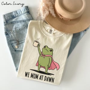 May include: An ivory t-shirt featuring a graphic of a green frog wearing a pink cape and holding a coffee mug, with the text "WE MOM AT DAWN" printed below. The shirt is paired with light blue denim jeans, a tan wide-brimmed hat, and a silver necklace.