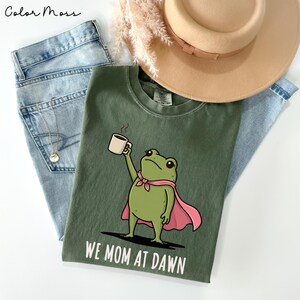 May include: A moss green t-shirt featuring a graphic of a green frog wearing a pink cape and holding a steaming mug. The text "WE MOM AT DAWN" is printed in white below the frog. The shirt is layered with light blue denim jeans and a tan hat.