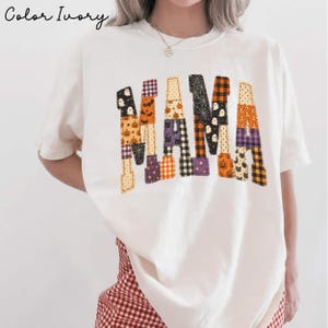 May include: An ivory t-shirt featuring the word MAMA printed in a patchwork style. The letters are composed of various Halloween-themed patterns including pumpkins, ghosts, spiderwebs, and plaid in orange, black, purple, and white colors.