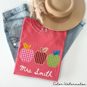 Op de afbeelding: Een watermeloenkleurig t-shirt met drie appelvormige patches met ruit- en streepmotieven, de tekst Mrs Smith in wit handschrift, gecombineerd met een lichtblauwe spijkerbroek, een beige hoed met brede rand en een fijne gouden ketting.