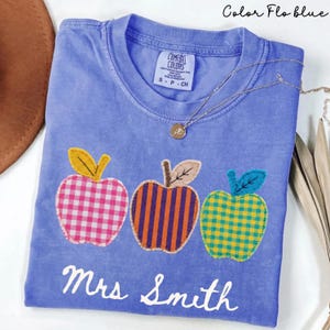 May include: A periwinkle blue short-sleeve t-shirt featuring three embroidered apple appliques. The left apple is pink and white gingham, the centre is orange and purple striped, and the right is yellow and teal gingham. The text Mrs Smith is printed in white.