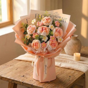 May include: A bouquet of handmade crochet flowers in shades of peach, pink, and white, wrapped in light pink paper. The bouquet includes roses and smaller accent flowers, with greenery. The words "FLOWER ART" are printed on the paper.