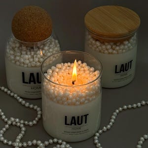 May include: Three glass candle jars filled with white wax and topped with small white pearls. One candle is lit with a visible flame. Each jar features a black label with the text LAUT HOME. One jar has a cork lid, one has a bamboo lid.