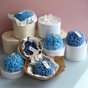 May include: A collection of round, white gift boxes containing blue, flower-shaped candles. Each candle features a small, rectangular white tag with the text LAUT printed on it. Some candles are nestled in crinkled white or brown paper liners.