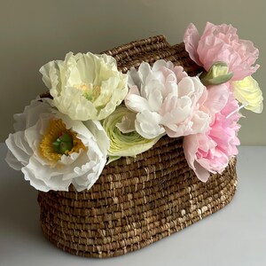 May include: A woven brown oval basket filled with an arrangement of artificial flowers. The bouquet features large white, pale yellow, and soft pink blossoms with layered petals, including some with prominent yellow centres and green accents.
