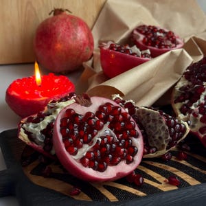 May include: A whole pomegranate, several pomegranate segments, and a red pomegranate-shaped candle with a lit flame. The fruit pieces reveal deep red arils and white membranes, arranged on a dark wooden board with a striped pattern.