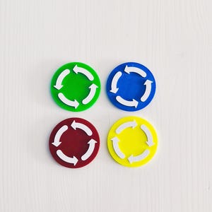 Könnte beinhalten: Vier kreisförmige Scheiben in Grün, Blau, Dunkelrot und Gelb, die jeweils ein weißes, geprägtes Recycling-Symbol mit drei in einer durchgehenden Schleife angeordneten Pfeilen aufweisen. Die Scheiben sind in einem zwei-mal-zwei Raster angeordnet.
