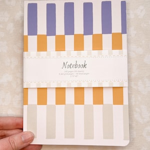 May include: A notebook with a striped design in lavender, orange, and cream. The notebook is held by a hand and has the word "Notebook" printed on the cover. The notebook is 5.75" x 8" and has 100 pages.