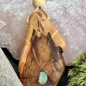 May include: A handcrafted wooden pendant with a natural, organic shape. The pendant features a light-coloured, polished wood grain with a dark, textured centre. An oval-shaped opal is set into the wood. The pendant hangs from a beige leather cord with a wooden bead.