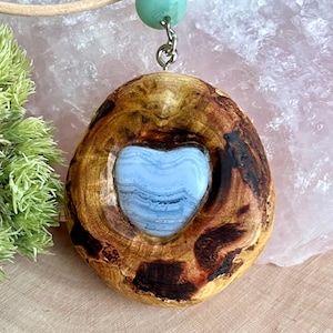 Puede incluir: Colgante de madera hecho a mano con una incrustación de piedra azul en forma de corazón. El colgante está suspendido de un cordón de cuero y presenta una pequeña cuenta turquesa redonda. La madera es de color marrón cálido natural con acentos marrón oscuro.