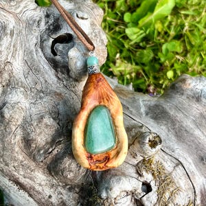 Könnte beinhalten: Ein Anhänger mit einem glatten, polierten grünen Stein in einem natürlichen, hellbraunen Holzrahmen. Das Schmuckstück enthält eine kleine, runde, hellgrüne Perle über dem Holz und hängt an einer dünnen, braunen Kordelkette.
