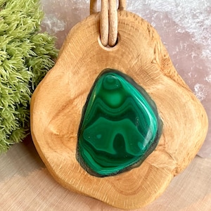 Könnte beinhalten: Handgefertigter Holzanhänger mit einem leuchtend grünen Malachit-Steineinsatz. Der Anhänger ist an einer natürlichen Lederschnur befestigt. Die Holzmaserung ist sichtbar und verleiht diesem einzigartigen Schmuckstück einen rustikalen Charme.