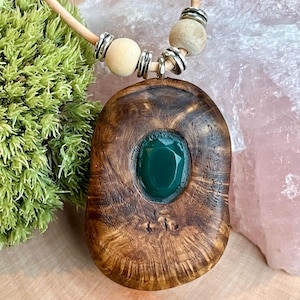 Könnte beinhalten: Eine handgefertigte Holz-Anhänger-Halskette mit einem dunkelbraunen, ovalen Anhänger. Der Anhänger hat einen leuchtend grünen Edelstein in der Mitte. Die Halskette hat eine hellbraune Lederschnur, Holzperlen und silberfarbene Metallakzente.