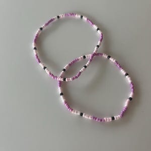 Puede incluir: Dos pulseras de cuentas. Cada pulsera está hecha de pequeñas cuentas redondas en tonos rosa, blanco y negro. Las pulseras son circulares y descansan sobre una superficie gris claro. Las cuentas están ensartadas para formar una banda flexible.