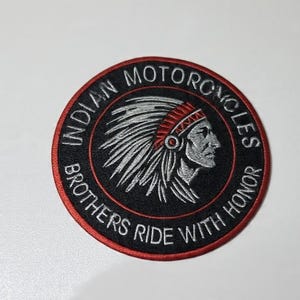 May include: A circular embroidered patch featuring a black background with a red border. The center displays a silver-toned profile of a person wearing a feathered headdress. The text reads INDIAN MOTORCYCLES BROTHERS RIDE WITH HONOR.
