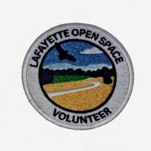 May include: A circular embroidered patch featuring a landscape scene with a golden field, a winding path, green trees, and a dark bird silhouette against a blue sky. The text LAFAYETTE OPEN SPACE is arched at the top and VOLUNTEER is at the bottom.