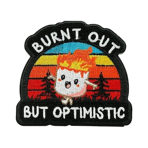 May include: Embroidered patch with the words "BURNT OUT" above and "BUT OPTIMISTIC" below a cartoon marshmallow on fire. The marshmallow has a smiling face and is set against a sunset background with trees.