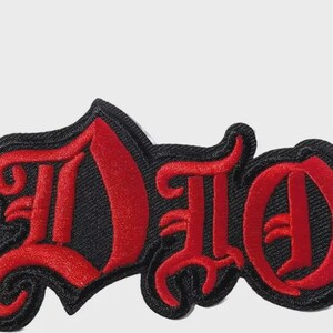 May include: An embroidered patch featuring the word DIO in a stylized red gothic font with a thick black border. The letters are stitched with a textured red thread against a black fabric background, creating a bold and high-contrast design.