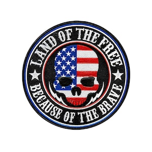 May include: A circular embroidered patch featuring a central skull graphic with a red, white, and blue American flag pattern. The black border contains the white text LAND OF THE FREE and BECAUSE OF THE BRAVE separated by two white stars.