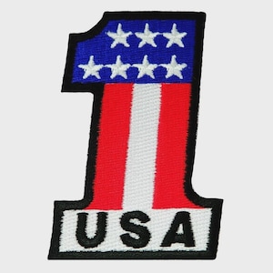 May include: An embroidered patch in the shape of the number one. The top section is blue with seven white stars. The vertical body features red and white stripes. The base is white with the black text USA, all outlined in a thick black border.