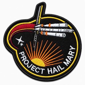Puede incluir: Un parche bordado circular con fondo negro que muestra una nave espacial blanca y naranja, una estela roja, un sol amarillo y una estrella blanca de cuatro puntas. El texto PROJECT HAIL MARY está bordado en blanco en el borde inferior.
