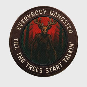 Könnte beinhalten: Ein runder bestickter Aufnäher mit einer dunklen, geweihartigen Kreatur in einem rot-schwarzen Wald. Der braune Rand enthält den weißen gestickten Text: EVERYBODY GANGSTER TILL THE TREES START TALKIN'. Das Design verwendet dunkle Farbtöne.