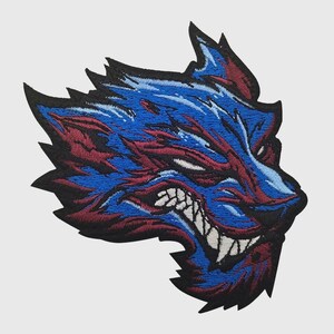 Könnte beinhalten: Ein gestickter Aufnäher mit einem stilisierten Wolfskopf im Profil. Das Design verwendet leuchtend blaue und dunkelrote Fäden mit schwarzen Umrandungen und zeigt einen grimmigen Ausdruck mit scharfen weißen Zähnen sowie gezackten, spitzen Fellstrukturen.
