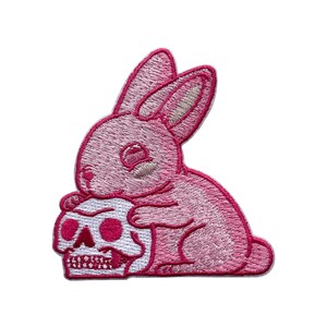 May include: Embroidered patch featuring a pink bunny hugging a white skull with pink accents. The bunny has detailed stitching and a cute expression. The skull has detailed teeth and eye sockets. This patch is suitable for jackets, bags, or other fabric items.