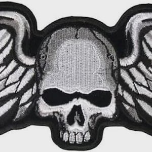 May include: An embroidered patch featuring a central human skull in shades of gray and white, flanked by two large, detailed wings extending horizontally. The entire design is outlined with a thick black border, creating a high-contrast graphic.