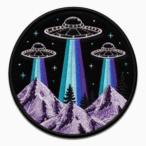 Könnte beinhalten: Ein runder Stickaufnäher mit drei silbernen UFOs, die über violetten Bergen und dunklen Tannen schweben. Jedes Raumschiff strahlt einen vertikalen Lichtstrahl in Blau- und Violetttönen auf einen schwarzen, mit kleinen Sternen verzierten Hintergrund.