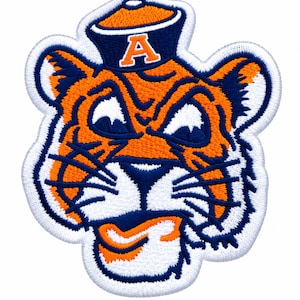 May include: An embroidered patch featuring a stylized tiger head with orange fur, dark blue stripes, and white accents. The tiger wears a dark blue sailor hat with an orange letter A on the front. The entire design is outlined in white thread.