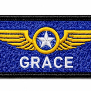 Op de afbeelding: Een rechthoekige geborduurde patch met een blauwe achtergrond en een zwarte rand. Het ontwerp toont een geel gevleugeld embleem met een witte ster in een gele cirkel. De naam GRACE is in witte hoofdletters onder het embleem geborduurd.