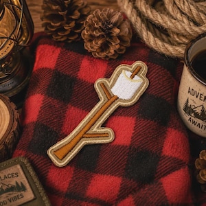 May include: An embroidered patch featuring a white marshmallow toasted to a golden brown, skewered on a brown wooden stick. The patch has a tan border and is shaped like a branch with a small twig extending from the side of the stick.