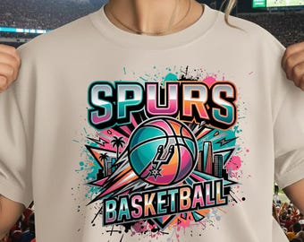 Könnte beinhalten: Beiges T-Shirt mit farbenfrohem Grafikdesign. Das Design zeigt das Wort "SPURS" in Türkis und Pink, einen Basketball mit dem Spurs-Logo und das Wort "BASKETBALL". Das Design zeigt auch Palmen und Stadtlandschaften.