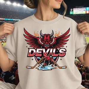 Könnte beinhalten: Beiges T-Shirt mit roter Teufelsgrafik mit Flügeln, Hörnern und dem Wort "DEVILS" in Weiß. Darunter eine Hockey-Spieler-Illustration mit gekreuzten Hockeyschlägern. Das Design ist mittig platziert.