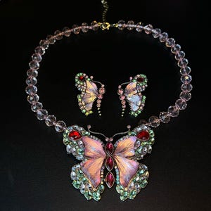 May include: A butterfly-themed jewellery set featuring a necklace and matching earrings. The necklace and earrings have a pink and purple butterfly design with red and green accents. The necklace is made of clear, pink beads.
