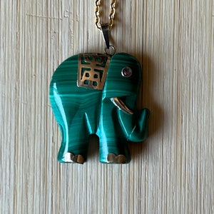 May include: A green malachite elephant pendant with gold accents and a pink stone in the eye, hanging from a gold chain.