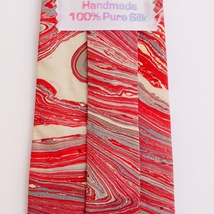 May include: A red and cream silk tie with a marbled pattern. The tie has a label that reads "Handmade 100% Pure Silk".