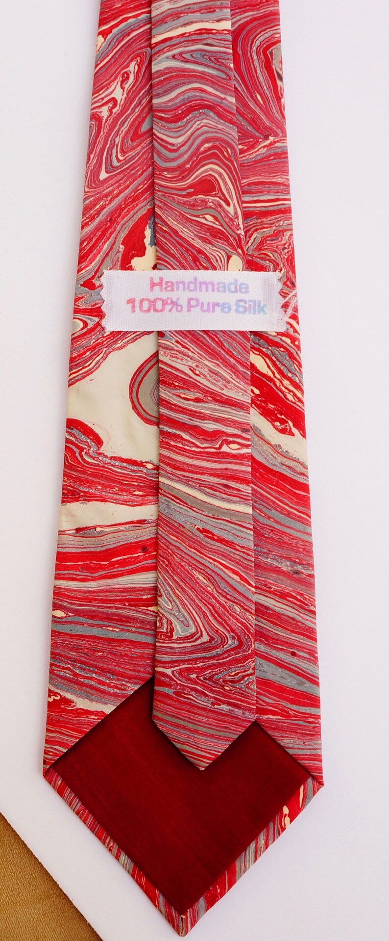 May include: A red and cream silk tie with a marbled pattern. The tie has a label that reads "Handmade 100% Pure Silk".