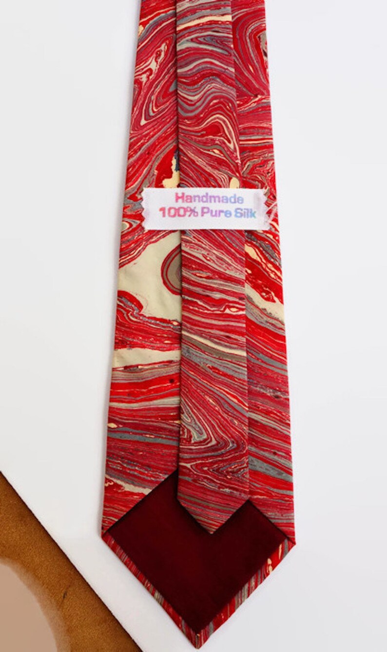 May include: A red and cream silk tie with a marbled pattern. The tie has a burgundy lining and a label that reads "Handmade 100% Pure Silk".